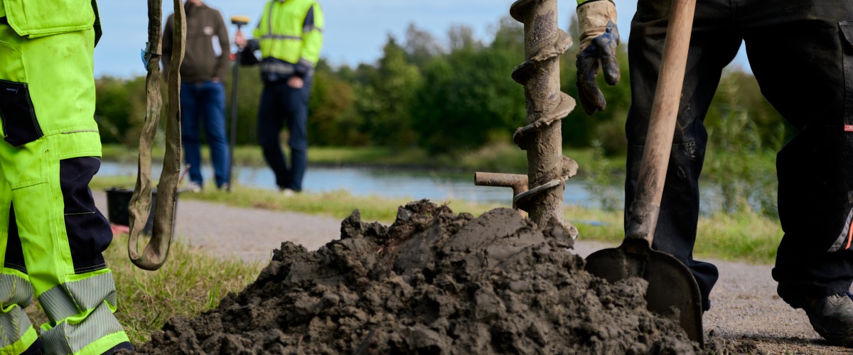 Im Vordergrund ist ein Haufen brauner Erde zu sehen, in dem eine Schaufel und ein Bohrgestänge stecken.Links und rechts davon stehen Menschen, die Sicherheitskleidung tragen. Im Hintergrund sind ebenfalls zwei Personen zu erkennen, die das Geschehen aus der Entfernung zu beobachten scheinen.