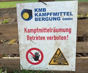 Auf Holzplanken angebrachtes Schild mit der Aufschrift "KMB Kampfmittelbergung GmbH - Kampfmittelräumung: Betreten verboten!" Unter der Schrift sind zwei Zeichen abgebildet: Eins zeigt einen roten Kreis, der in der Mitte durchgestrichen ist, dahinter befindet sich das Handsymbol. Das andere Zeichen ist ein gelbes Dreieck, das schwarz umrandet ist und aus dem Zentrum gehen schwarze Striche ab, was eine Explosion symbolisieren soll.