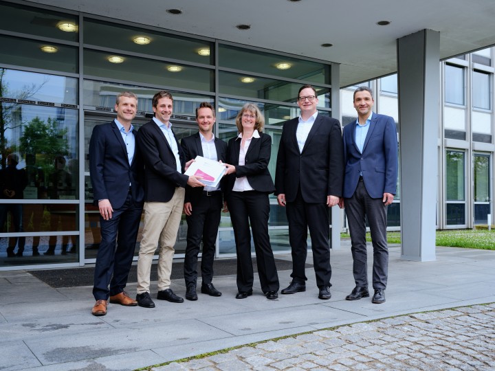 Sechs Menschen stehen nebeneinander vor einem gläsernen Eingang, ein Mann überreicht einer Frau einen Ordner. Alle lachen.