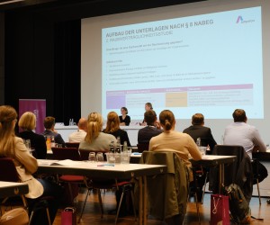 Vortragssituation: Auf einer Leinwand ist eine Präsentation mit Informationen zu sehen, vor der Leinwand stehem zwei Personen an Brückentischen. Noch weiter im Vordergrund sind einige Tischreihen zu sehen, an denen Menschen sitzen, die Richtung Leinwand blicken.