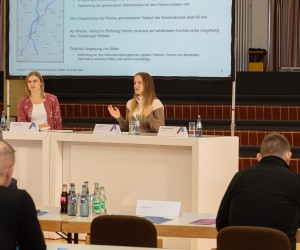 Informationsveranstaltung für Täger öffentlicher Belange am 11. November 2024: Zu sehen sind zwei Frauen, die hinter zwei großen weißen Tischen vor einer Leinwand stehen. Mehrere Tischreihen sind im Vordergrund aufgebaut, daran sitzen etwaige Personen mit dem Rücken zur Kamera.