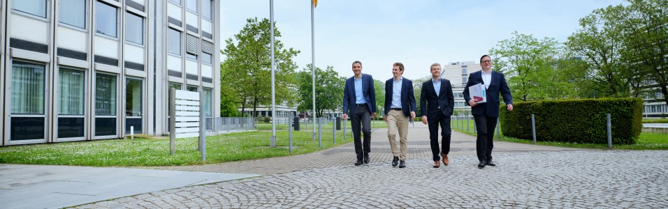 Vier Personen laufen nebeneinander Richtung Kamera, einer hält einen Ordner in der Hand.