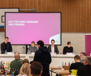 Informationsveranstaltung für Träger öffentlicher Belange am 25. Januar 2025: Zu sehen sind vier Personen, die hinter drei großen weißen Tischen vor einer Leinwand stehen. Mehrere Tischreihen sind im Vordergrund aufgebaut, daran sitzen etwaige Personen mit dem Rücken zur Kamera. Zwischen den Tischreihen steht eine Person, die einer anderen sitzenden Person ein Mikrofon hinhält.