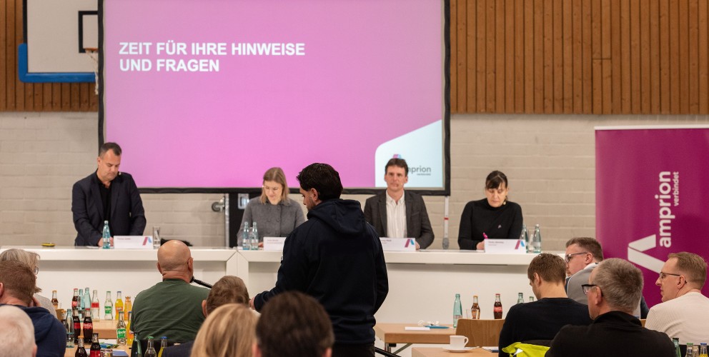 Informationsveranstaltung für Träger öffentlicher Belange am 25. Januar 2025: Zu sehen sind vier Personen, die hinter drei großen weißen Tischen vor einer Leinwand stehen. Mehrere Tischreihen sind im Vordergrund aufgebaut, daran sitzen etwaige Personen mit dem Rücken zur Kamera. Zwischen den Tischreihen steht eine Person, die einer anderen sitzenden Person ein Mikrofon hinhält.
