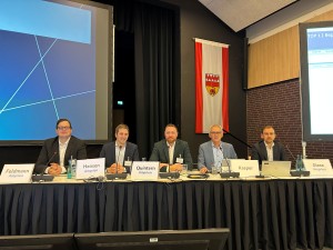 Fünf Männer im Anzug sitzen an einem langen Tisch, der vorne mit Stoff verkleidet ist. Auf dem Tisch stehen Namensschilder. Alle blicken in Richtung Kameralinse.