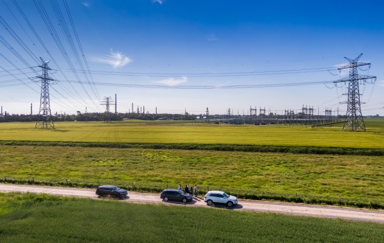 Landschaftsbild: Auf der Straße vorne stehen drei Autos, im Hintergrund sind eine Industrianlage sowie Freileitungen zu sehen.